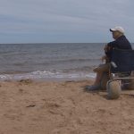 Man sitting at water's edge next to a wheelchair with wide wheels